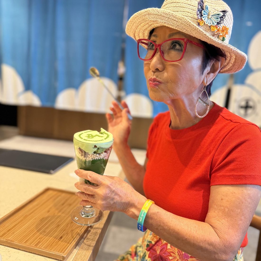 Olelo paʻa in a red top and red glasses, playfully sipping a green smoothie