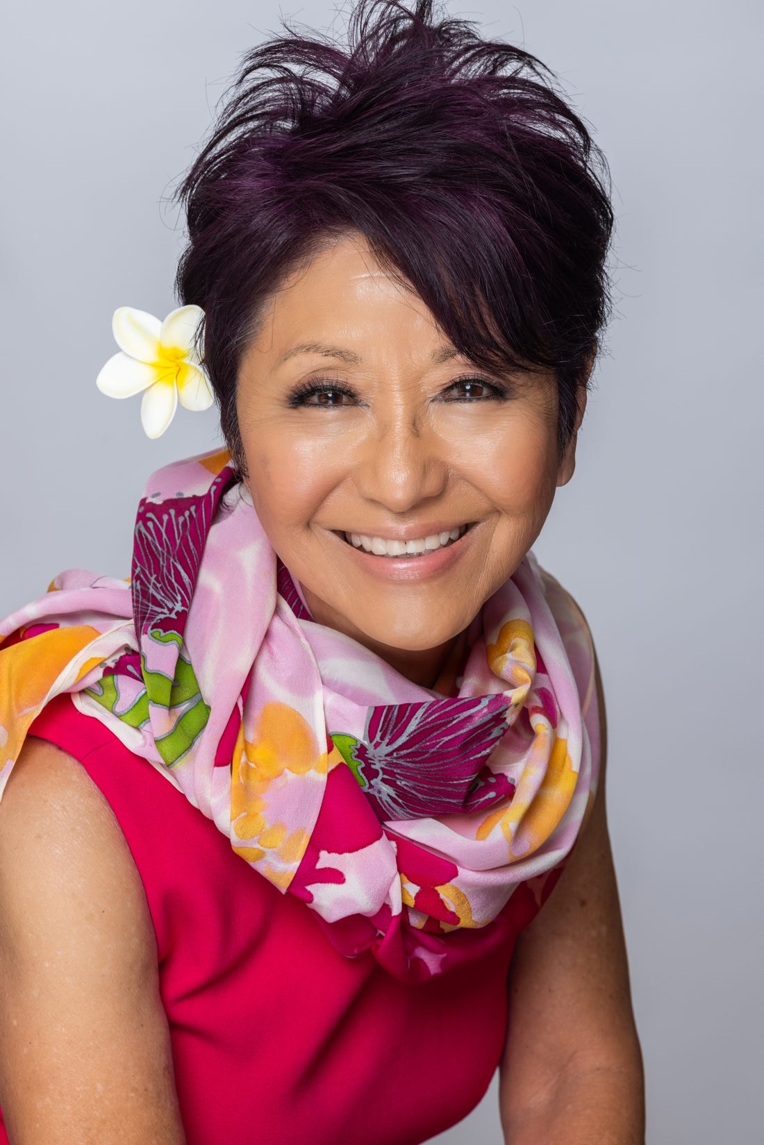 Olelo paʻa Ogawa smiling in a pink dress and floral scarf with a plumeria flower in her hair