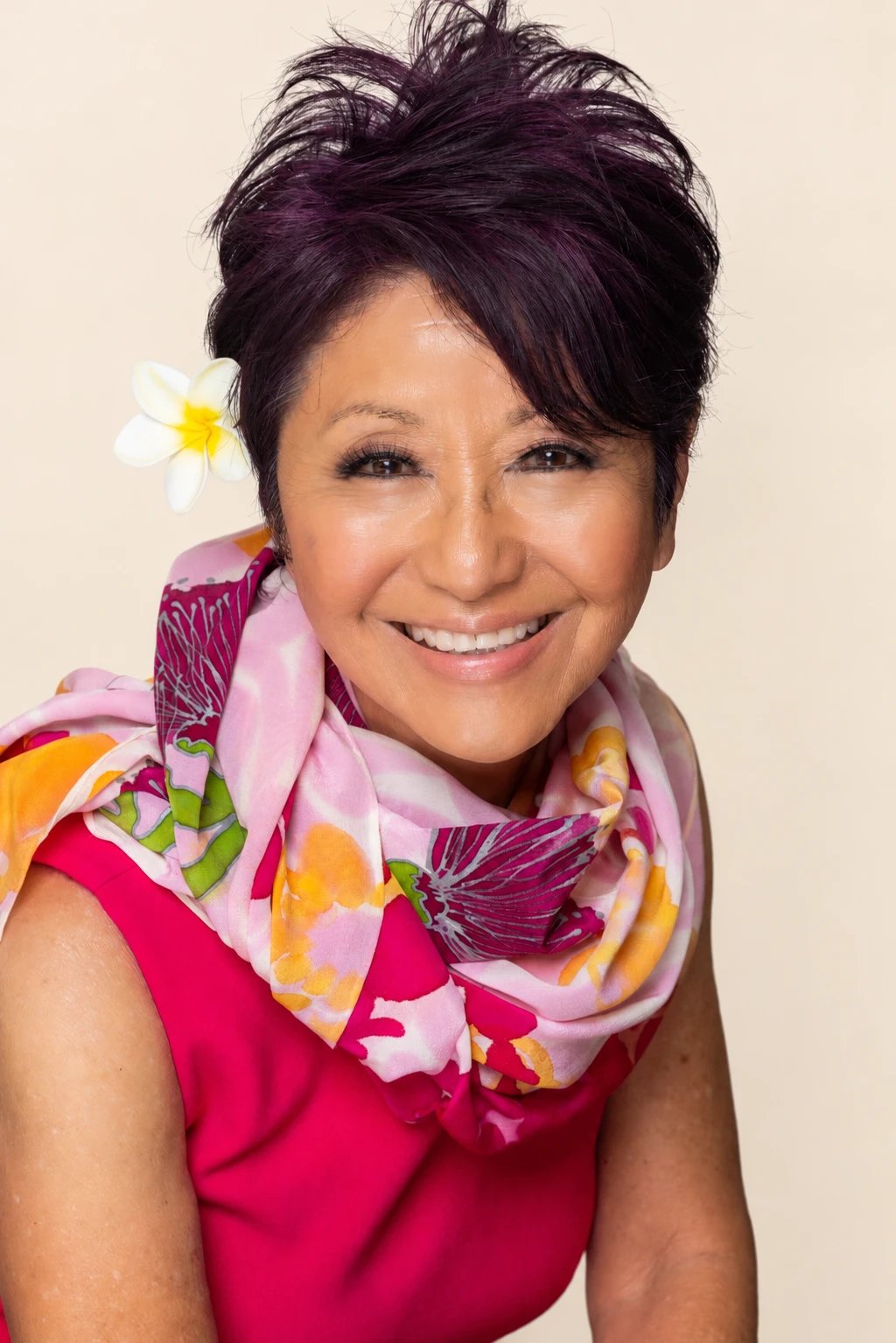 Olelo paʻa Ogawa smiling in a pink dress and floral scarf with a plumeria flower in her hair