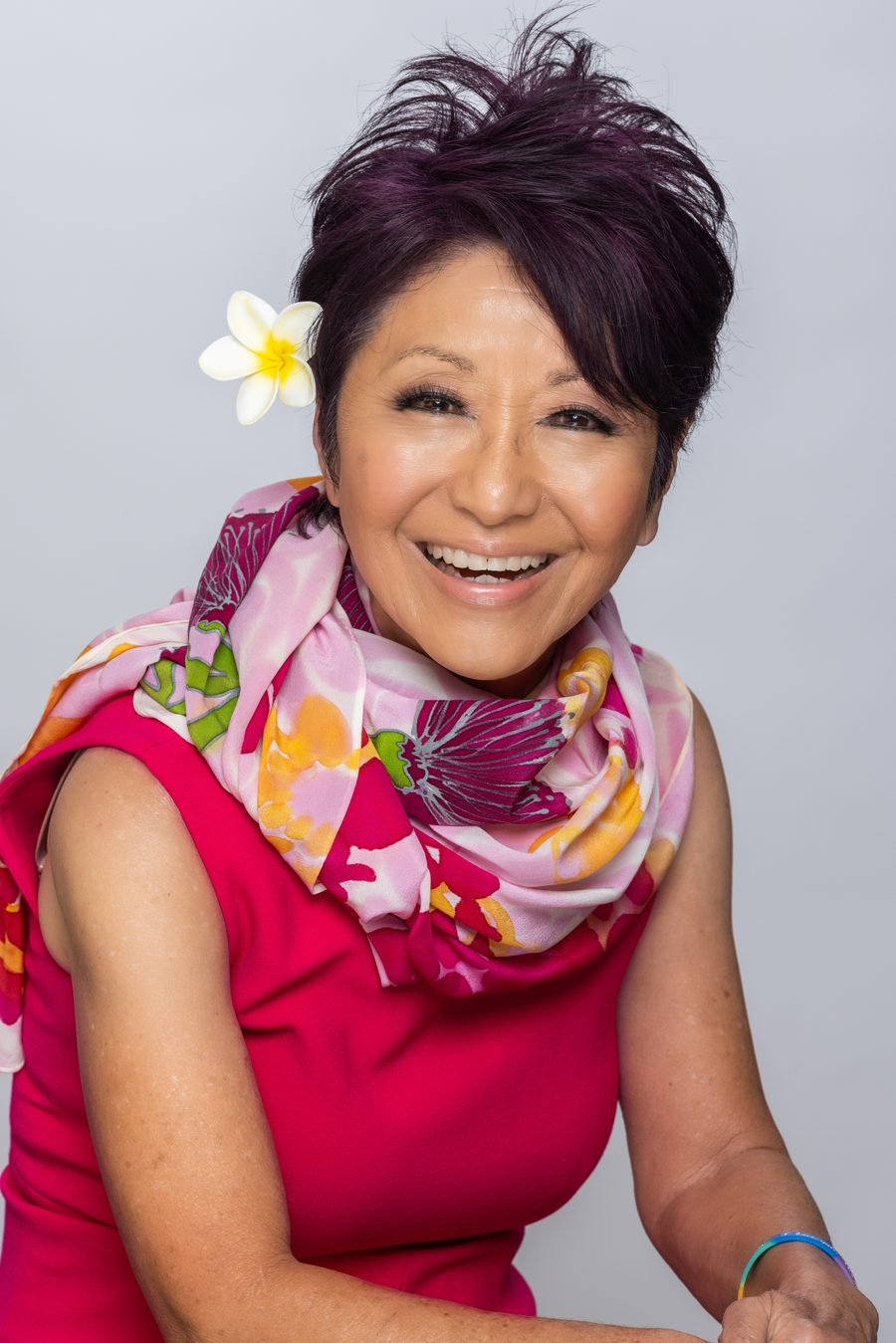 Olelo paʻa laughing with her arms crossed, in a bright pink dress and floral scarf with a plumeria in her hair